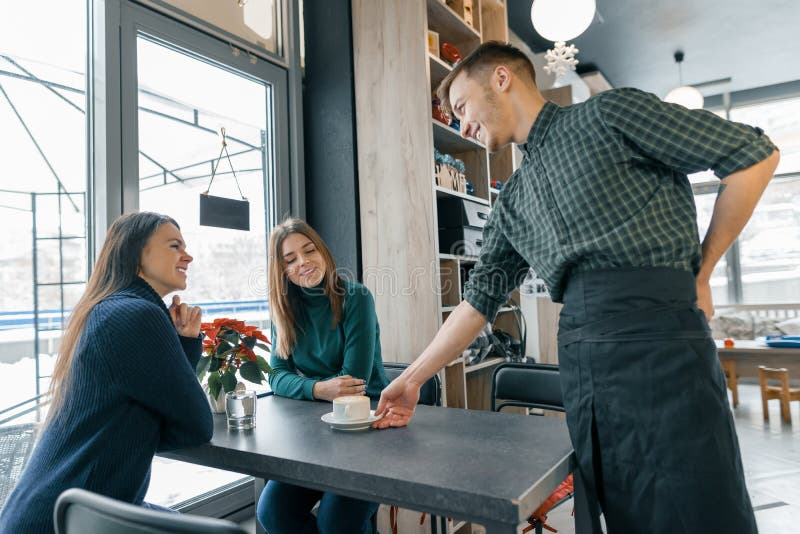 Café, saison d'hiver, deux jeunes femmes s'asseyant à la table, et un barman masculin avec une tasse de café souriant et parlant image stock Café, saison d'hiver, deux jeunes femmes s'asseyant à la table, et un barman masculin avec une tasse de café souriant et parlant image stock