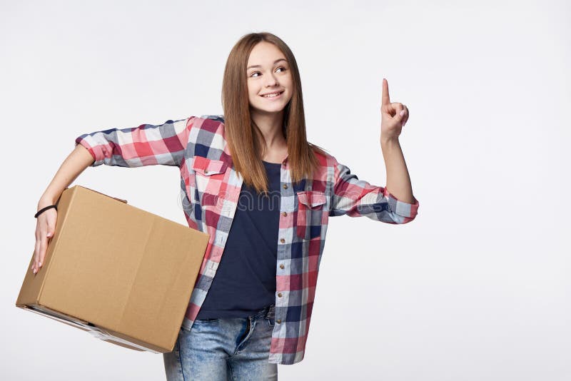 Delivery, relocation and unpacking. Smiling young woman holding cardboard box and pointing finger up or pushing imaginary button. Delivery, relocation and unpacking. Smiling young woman holding cardboard box and pointing finger up or pushing imaginary button