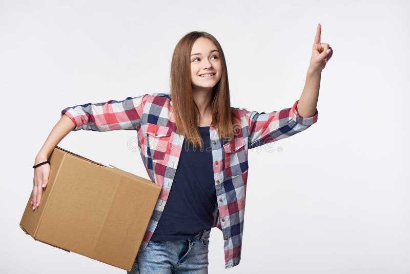 Delivery, relocation and unpacking. Smiling young woman holding cardboard box and pointing finger up or pushing imaginary button. Delivery, relocation and unpacking. Smiling young woman holding cardboard box and pointing finger up or pushing imaginary button