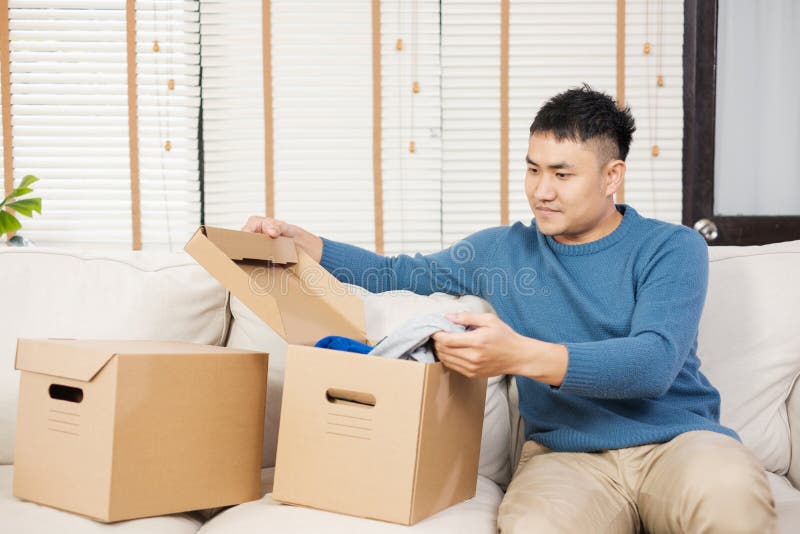 Cajas de cartón abiertas del hombre asiático mientras que se mueve al nuevo hogar en el sofá en sala de estar desempaque para la fotos de archivo Cajas de cartón abiertas del hombre asiático mientras que se mueve al nuevo hogar en el sofá en sala de estar desempaque para la fotos de archivo
