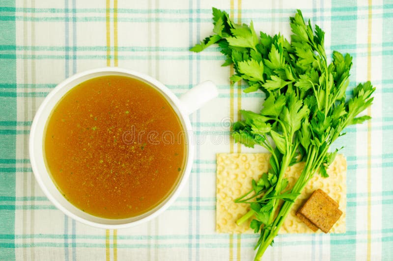 Bouillon, broth, clear soup in a white cup with a loaf, parsley, boiled egg on the tablecloth. Close-up. Bouillon, broth, clear soup in a white cup with a loaf, parsley, boiled egg on the tablecloth. Close-up.