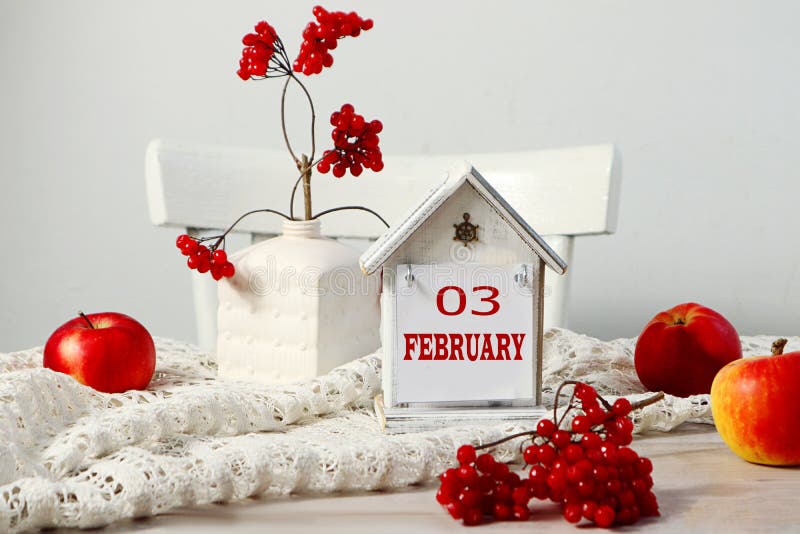 Calendar for February 3: the name of the month February in English, the numbers 03 on a decorative house among the branches of viburnum, red apples on a white napkin. Calendar for February 3: the name of the month February in English, the numbers 03 on a decorative house among the branches of viburnum, red apples on a white napkin.