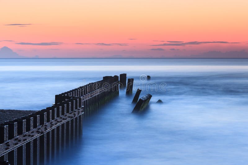 An unusually calm blue ocean at sunrise in Buxton, North Carolina where the coastal engineered groyne interrupts water flow of the Atlantic Ocean against the beach. An unusually calm blue ocean at sunrise in Buxton, North Carolina where the coastal engineered groyne interrupts water flow of the Atlantic Ocean against the beach.