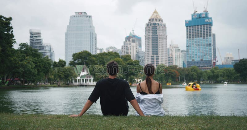 Calm Loving young man and woman sits by lake inside public city park with beautiful nature. Rear view boyfriend and