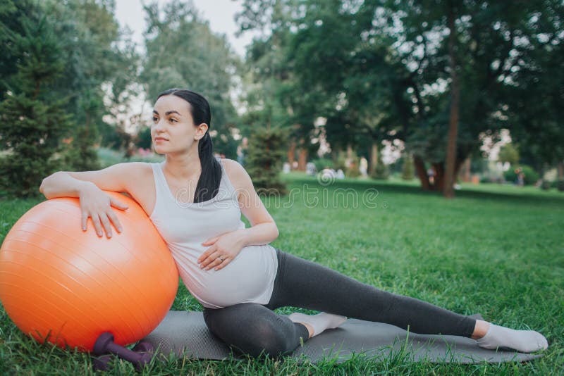 Calm and peacefyl young pregnant woman sit on yoga mate in park and look straight. She lean to orange fitness ball royalty free stock photography Calm and peacefyl young pregnant woman sit on yoga mate in park and look straight. She lean to orange fitness ball royalty free stock photography