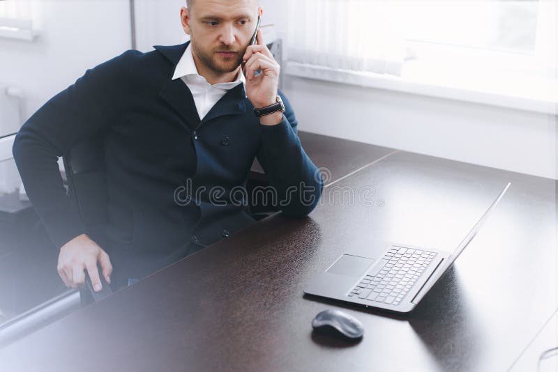Calm serious young man uses phone while working at table in office stock image Calm serious young man uses phone while working at table in office stock image