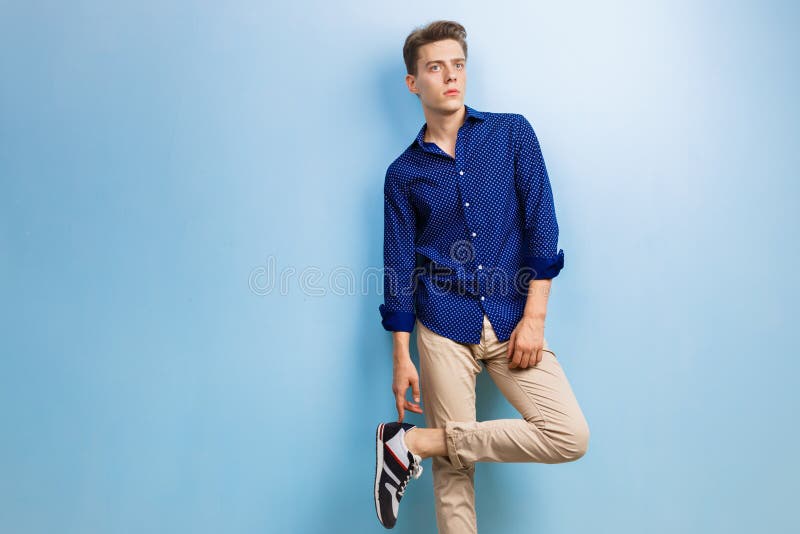 calm thoughtful handsome dark haired guy wearing blue shirt beige pants standing against wall 138679353
