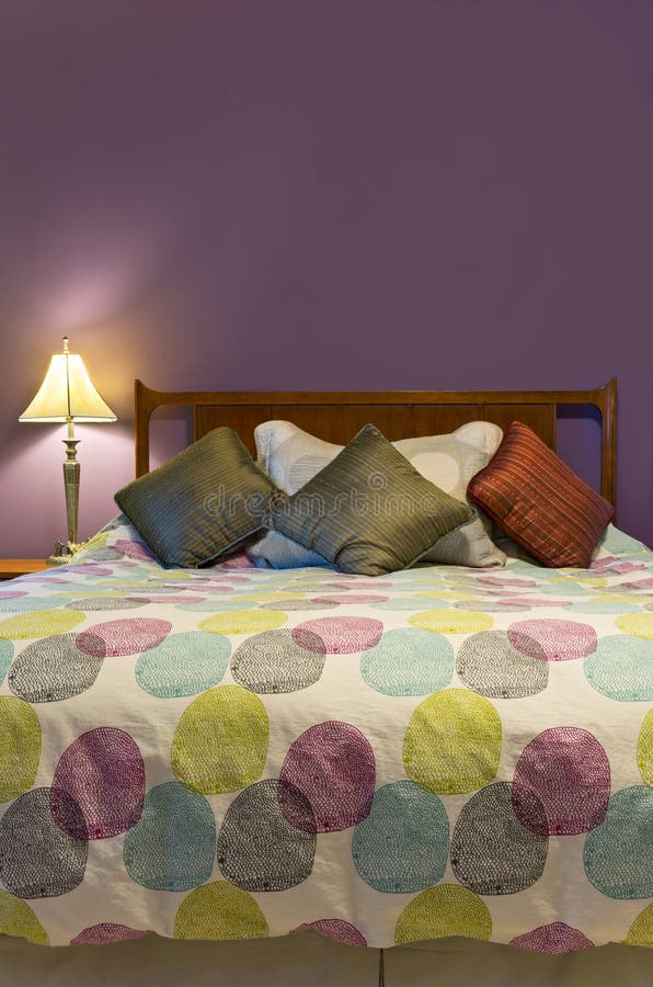 Bedroom interior with lamp colorful pillows and comforter decorating queen size bed and purple wall. Bedroom interior with lamp colorful pillows and comforter decorating queen size bed and purple wall