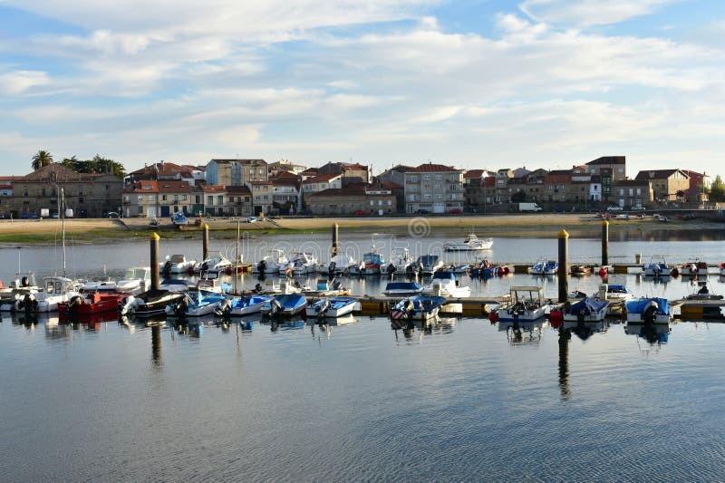 Small fishing village with pier and boats. Beach, harbour and promenade with trees. Sunset light, blue sky with clouds. Cambados