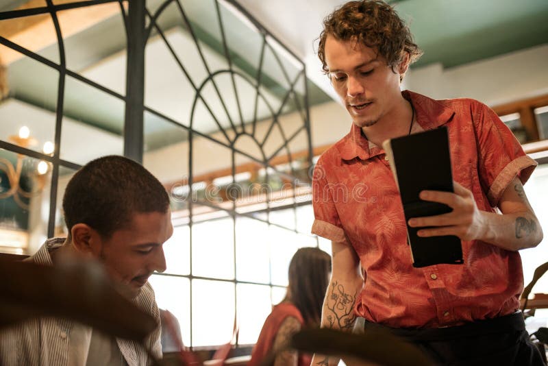 Young waiter taking an order from a young men reading a menu at a table in a trendy restaurant. Young waiter taking an order from a young men reading a menu at a table in a trendy restaurant