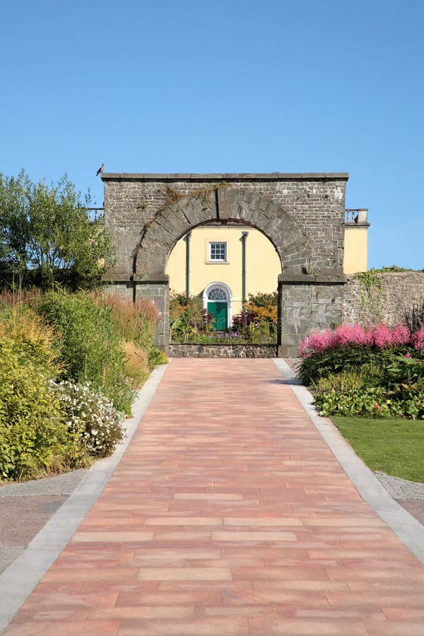 Terracotta path with flower borders leading to an old stone arch with a villa beyond, set against a blue sky. Terracotta path with flower borders leading to an old stone arch with a villa beyond, set against a blue sky.