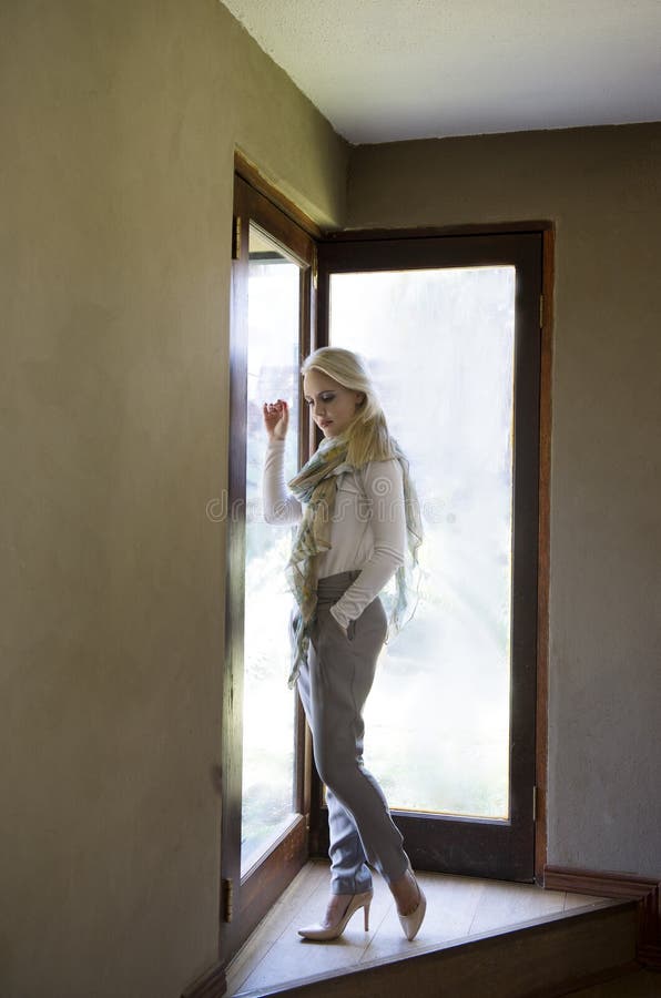 Camisa blanca que lleva de la mujer rubia sofisticada, pantalones grises, talones y bufanda, presentando al lado de las puertas d imágenes de archivo libres de regalías Camisa blanca que lleva de la mujer rubia sofisticada, pantalones grises, talones y bufanda, presentando al lado de las puertas d imágenes de archivo libres de regalías