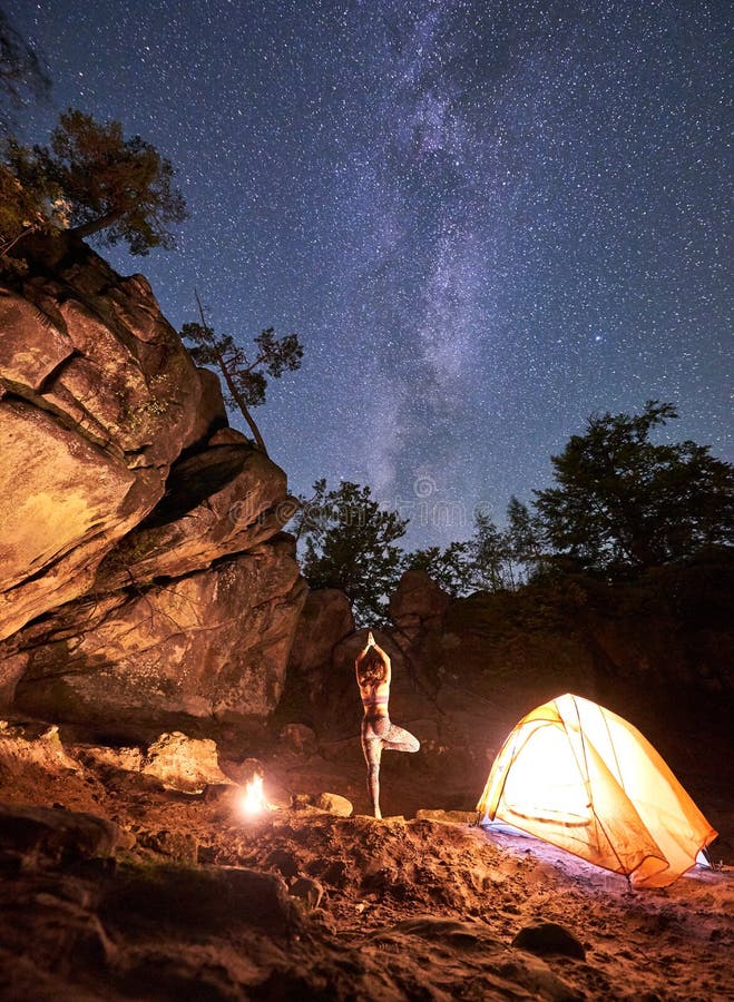 Night camping between mountains rocks. Lit by campfire back view silhouette of slim woman standing on one leg with raised arms doing yoga at tourist tent on clear starry sky background. Vrikshasana. Night camping between mountains rocks. Lit by campfire back view silhouette of slim woman standing on one leg with raised arms doing yoga at tourist tent on clear starry sky background. Vrikshasana