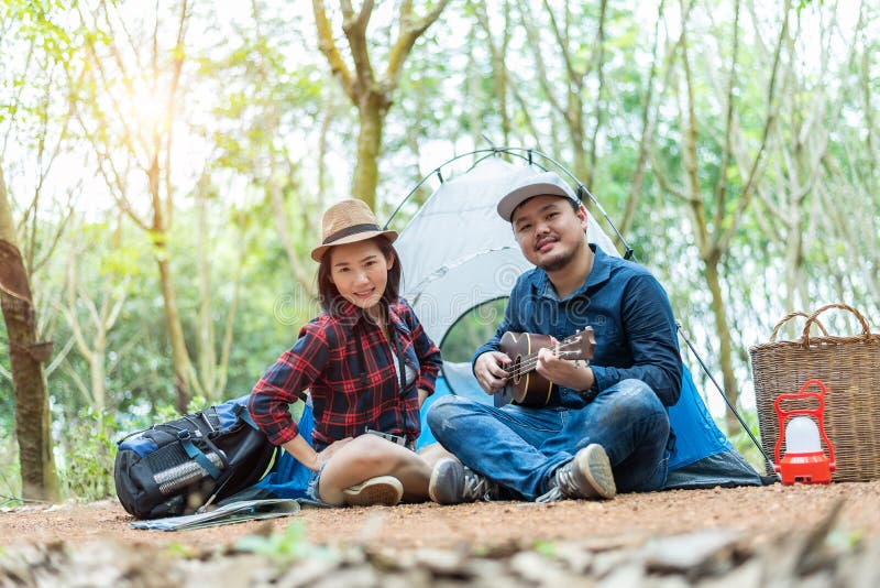 Camping asiatique de couples chez l'homme de forêt jouant l'ukulélé avec la femme devant la tente de camping Lampe et élément de images stock Camping asiatique de couples chez l'homme de forêt jouant l'ukulélé avec la femme devant la tente de camping Lampe et élément de images stock