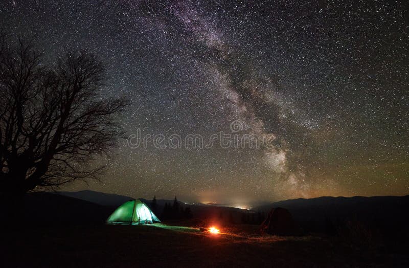 Bright bonfire burning near tourist illuminated tent. Night camping in mountain valley under beautiful sky full of stars and Milky way. Silhouette of big tree and distant hills in background. Bright bonfire burning near tourist illuminated tent. Night camping in mountain valley under beautiful sky full of stars and Milky way. Silhouette of big tree and distant hills in background