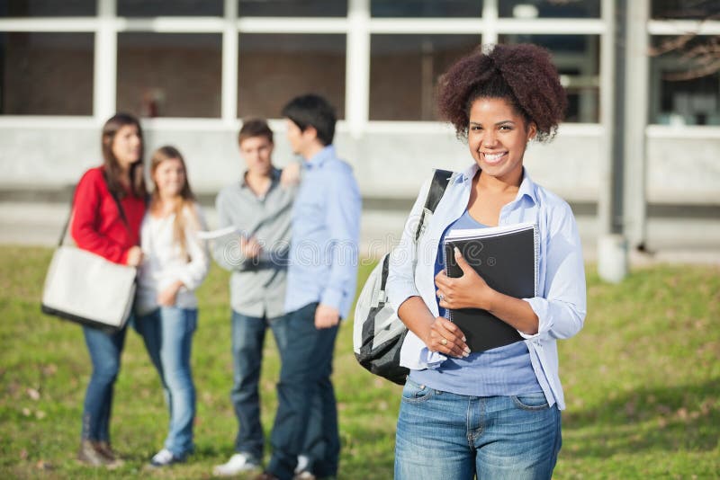 Portrait of confident female student holding books with friends standing in background on college campus. Portrait of confident female student holding books with friends standing in background on college campus