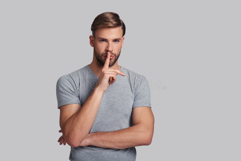 Thoughtful young man looking at camera and keeping index finger by his lips while standing against grey background. Thoughtful young man looking at camera and keeping index finger by his lips while standing against grey background