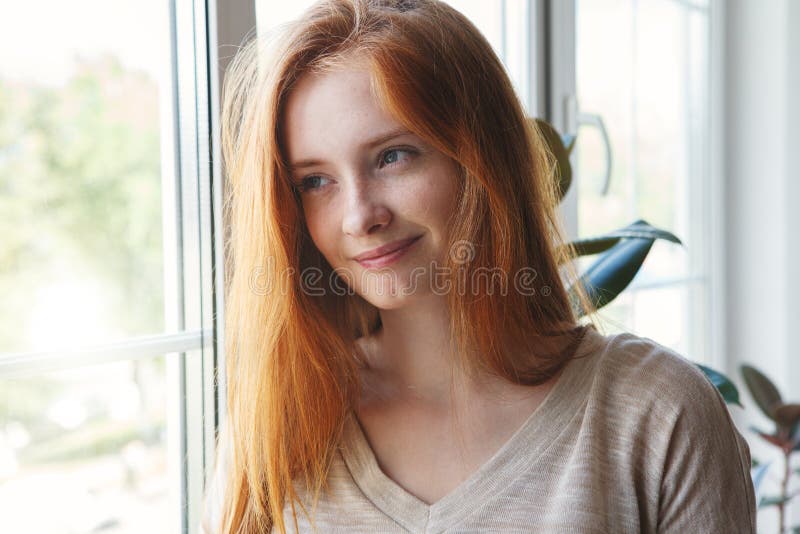 Indoor portrait of pretty smiling red haired young caucasian girl at home, sunny morning light