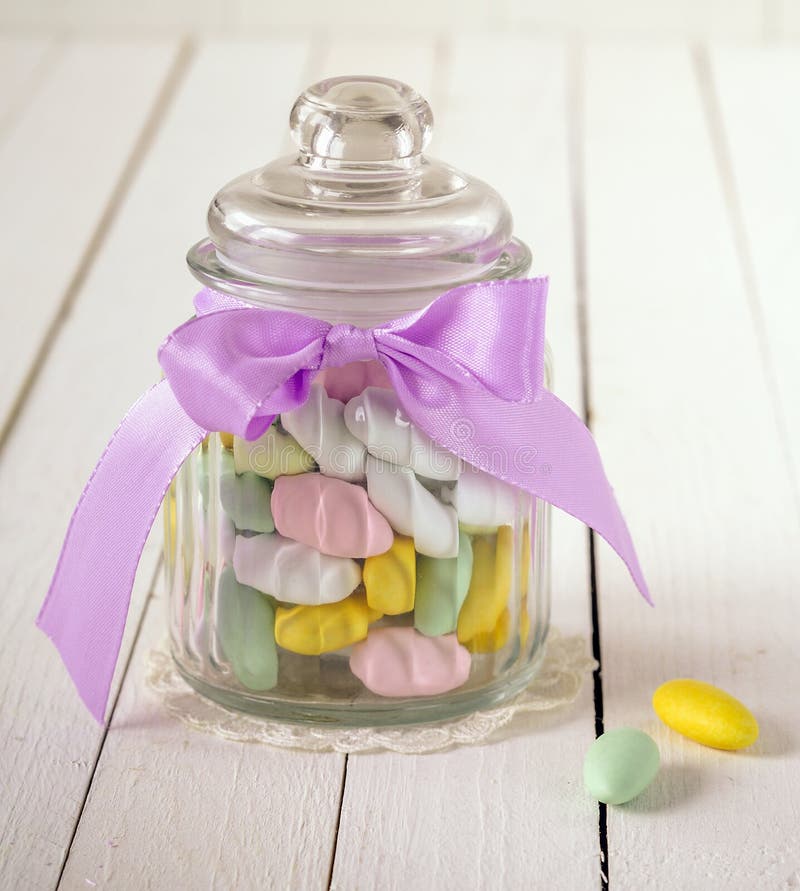 Close-up of an antique glass candy jar filled with colorful sugar covered almonds and a violet bow over a white wooden background. Close-up of an antique glass candy jar filled with colorful sugar covered almonds and a violet bow over a white wooden background