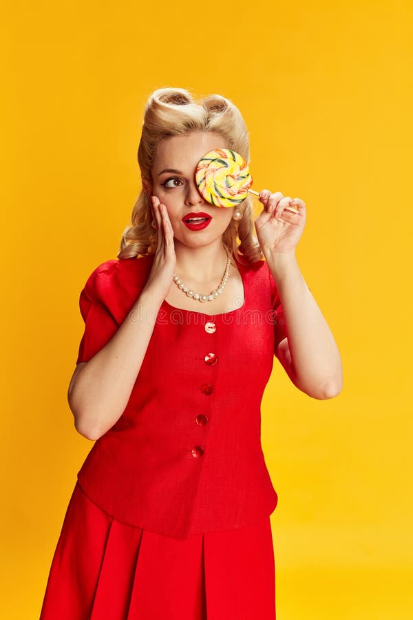 Candy lover. Portrait of beautiful young blonde girl with stylish hairstyle in red suit posing against yellow studio