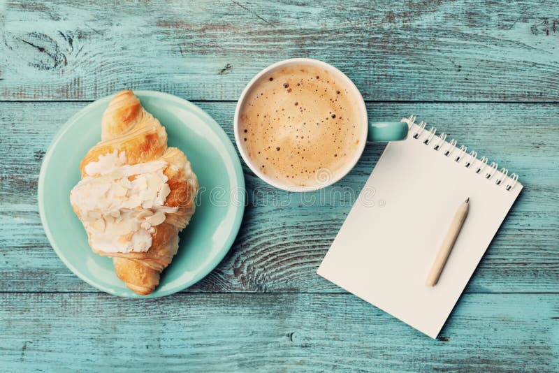 Caneca de café com croissant e caderno e lápis vazios para ideias do plano de negócios e do projeto na tabela rústica de turquesa