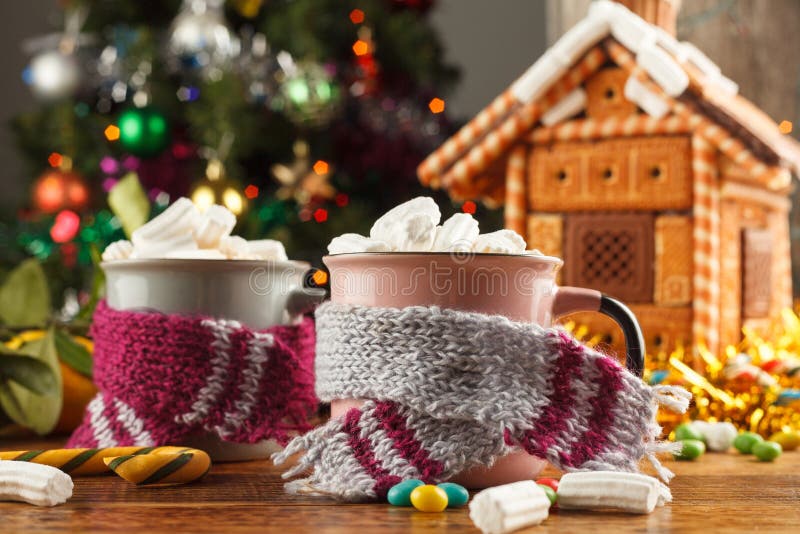 Canecas nos scarves com os marshmallows quentes e os doces de uma bebida no fundo de uma casa de pão-de-espécie Ainda vida festiv fotos de stock Canecas nos scarves com os marshmallows quentes e os doces de uma bebida no fundo de uma casa de pão-de-espécie Ainda vida festiv fotos de stock