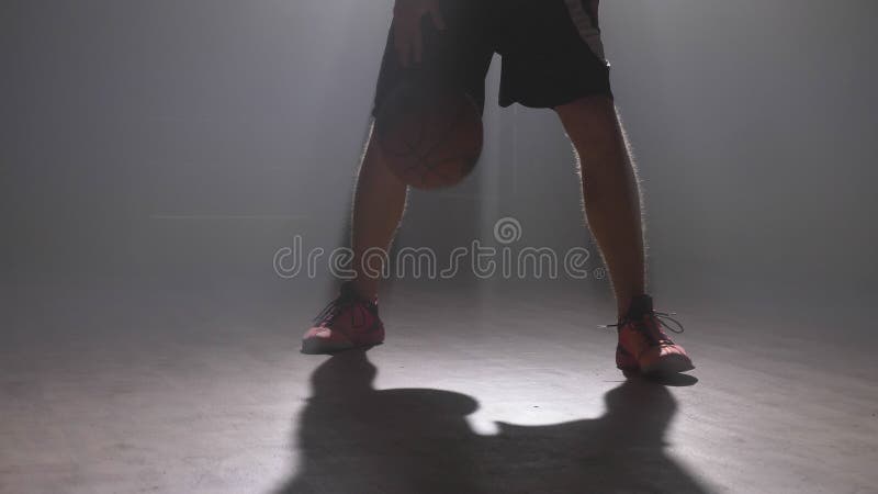 Cantidad cercana de las piernas del ` s del jugador de básquet que juegan con la bola en sitio brumoso oscuro con humo