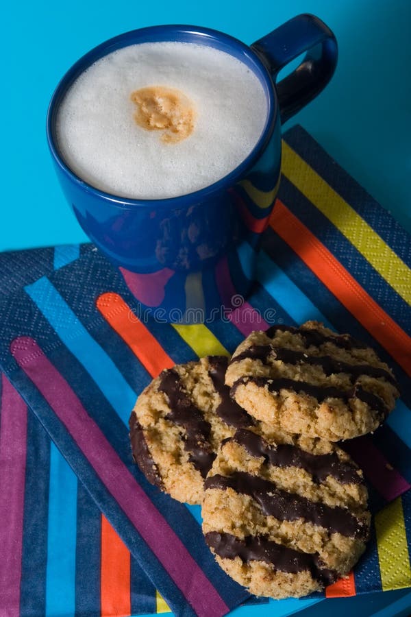 A blue cup of cappuccino with three chocolate biscuits and paper napkin. A blue cup of cappuccino with three chocolate biscuits and paper napkin