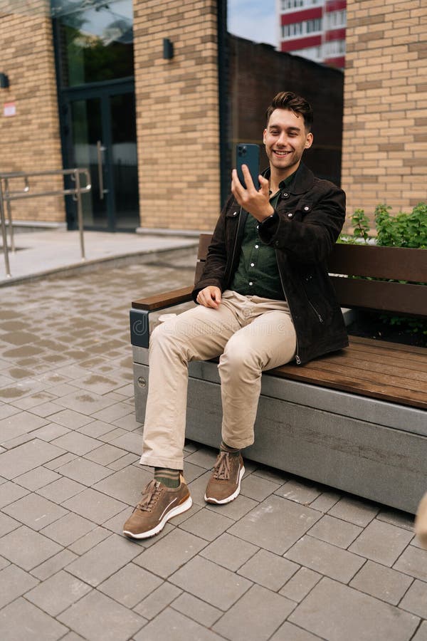 Capture verticale pleine longueur d'un homme positif en vêtements décontractés ayant chat vidéo en ligne sur le smartphone assis s photos stock