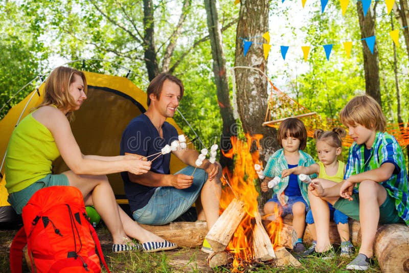 Big happy family, young parents and three kids, roasting marshmallows on the sticks over the fire at campsite in summer. Big happy family, young parents and three kids, roasting marshmallows on the sticks over the fire at campsite in summer