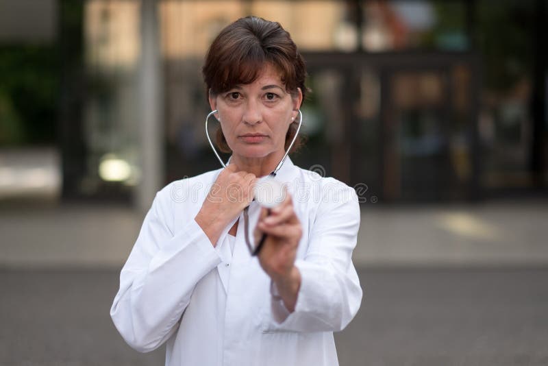 Pretty female cardiologist or doctor standing outdoors in front of the hospital holding a stethoscope with the disc extended as she prepares to do an examination and listen to the heart and lungs. Pretty female cardiologist or doctor standing outdoors in front of the hospital holding a stethoscope with the disc extended as she prepares to do an examination and listen to the heart and lungs