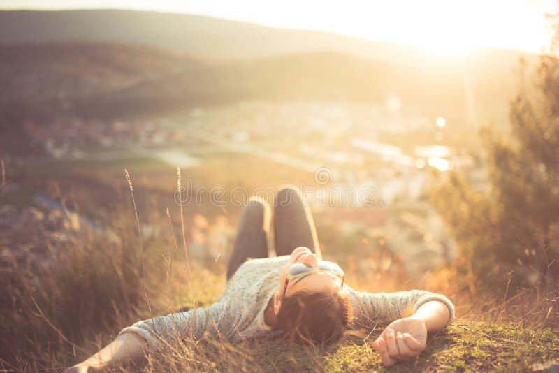 Carefree happy woman lying on green grass meadow on top of mountain edge cliff enjoying sun on her face. Enjoying nature sunset