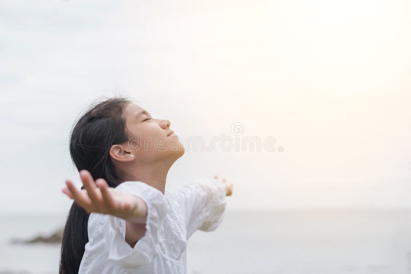 Carefree young Asian girl taking deep breath for natural blissful fresh air facing against sky, opening arms relaxing on the sea