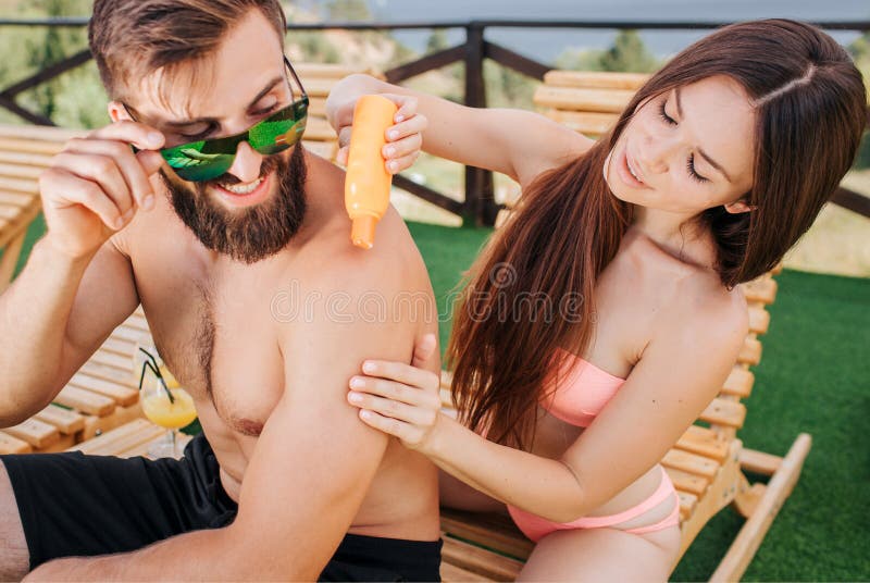 Careful girl sits behind guy and puts some sunproof cream on his shoulders. Also woman rubs them and holds orange bottle