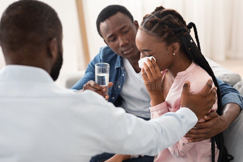 Caring Black Husband And Psychologist Comforting Crying Woman During Family Therapy Meeting At Marital Counselor`s Office, Loving Man Offering Glass Of Water To His Depressed Wife, Closeup