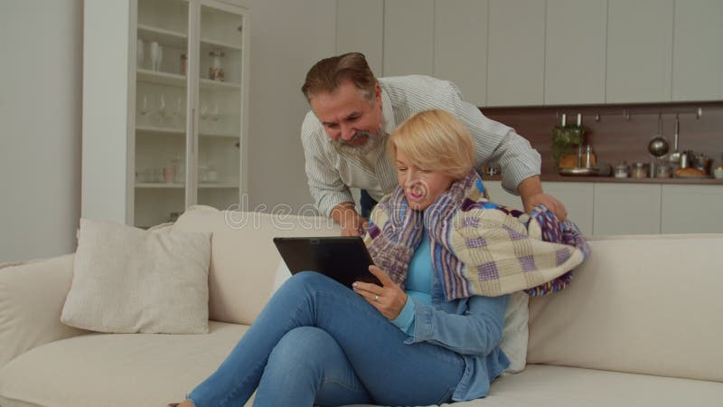 Caring handsome mature man covering lovely senior woman with blanket indoors