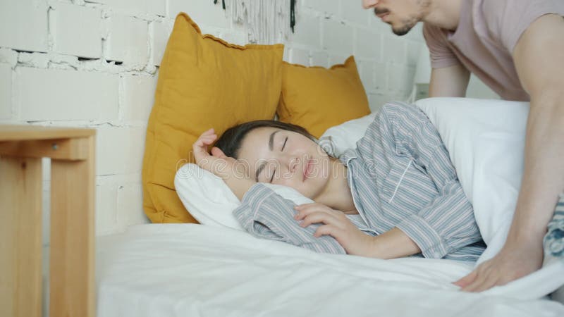 Caring husband covering sleeping wife with blanket and kissing in bedroom at home