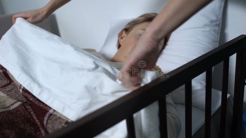 Caring nurse covering female patient who sleeping with teddy-bear with blanket