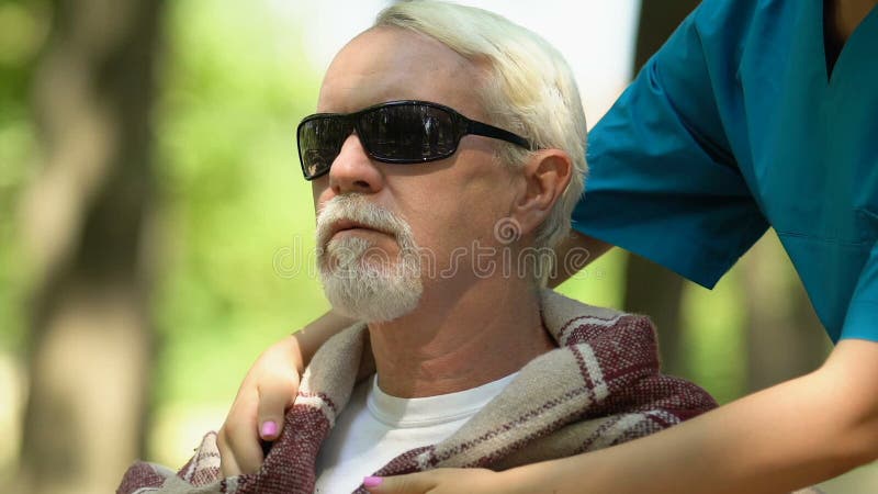 Caring nurse of nursing home covering shoulders of old blind man with plaid