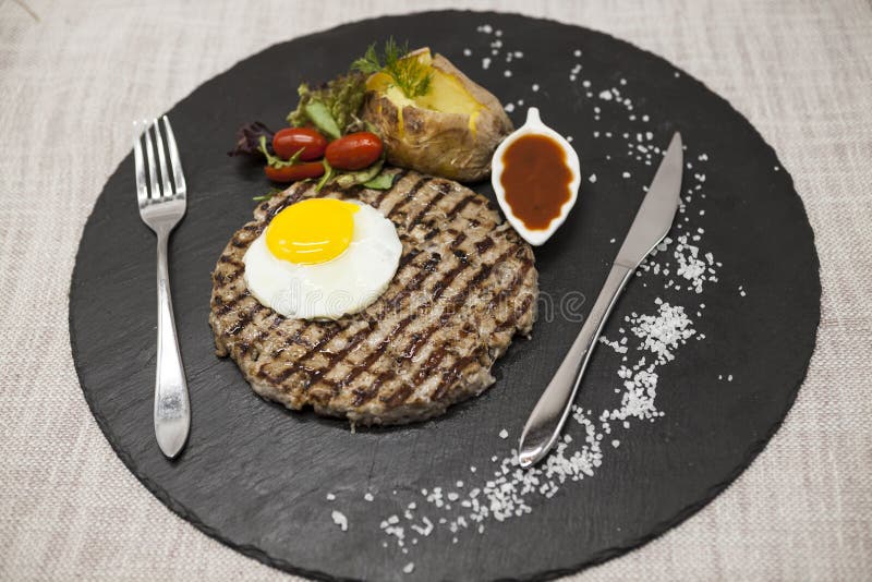 A carne marmoreada bife grelhada suculenta grande com ovo cozeu batatas com molho de assado Servido em uma placa de pedra com uma foto de stock royalty free A carne marmoreada bife grelhada suculenta grande com ovo cozeu batatas com molho de assado Servido em uma placa de pedra com uma foto de stock royalty free