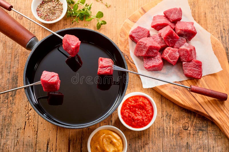 Tender prime beef being cooked in a fondue with cubed portions displayed on forks above the hot oil in the pot and herbs, spice and dips displayed to the side, overhead view. Tender prime beef being cooked in a fondue with cubed portions displayed on forks above the hot oil in the pot and herbs, spice and dips displayed to the side, overhead view