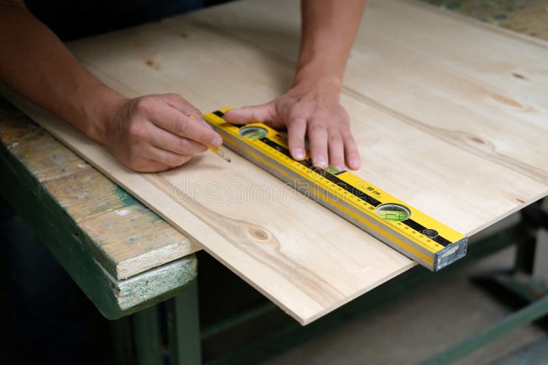Carpenter marking straight line on plywood sheet using spirit level in carpentry workshop. Measuring, drawing line, DIY concept.