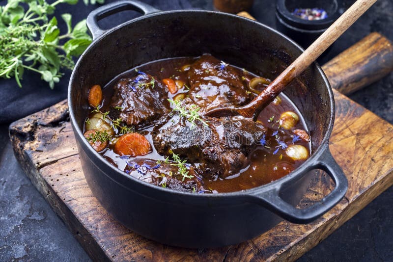 Traditional German braised beef cheeks in brown red wine sauce with carrots and onions offered as closeup in a cast iron Dutch oven on an old rustic cutting board. Traditional German braised beef cheeks in brown red wine sauce with carrots and onions offered as closeup in a cast iron Dutch oven on an old rustic cutting board