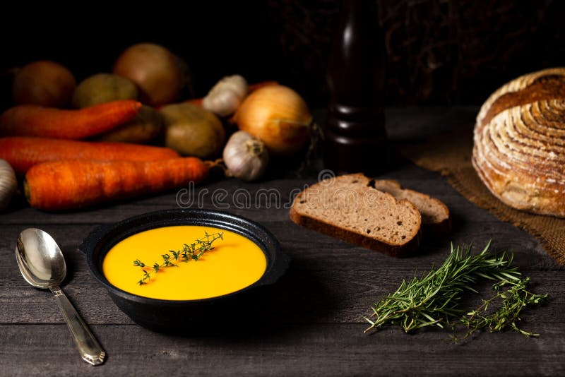 Carrot and potato soup in a black tureen with rosemary twig on a grey wood table, with a vintage silver spoon, onions, potatoes stock image Carrot and potato soup in a black tureen with rosemary twig on a grey wood table, with a vintage silver spoon, onions, potatoes stock image