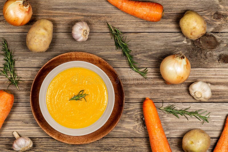 Carrot and potato soup bowl on drift wood table with a spread of vegetables, garlic, and rosemary. Flat lay royalty free stock photo Carrot and potato soup bowl on drift wood table with a spread of vegetables, garlic, and rosemary. Flat lay royalty free stock photo