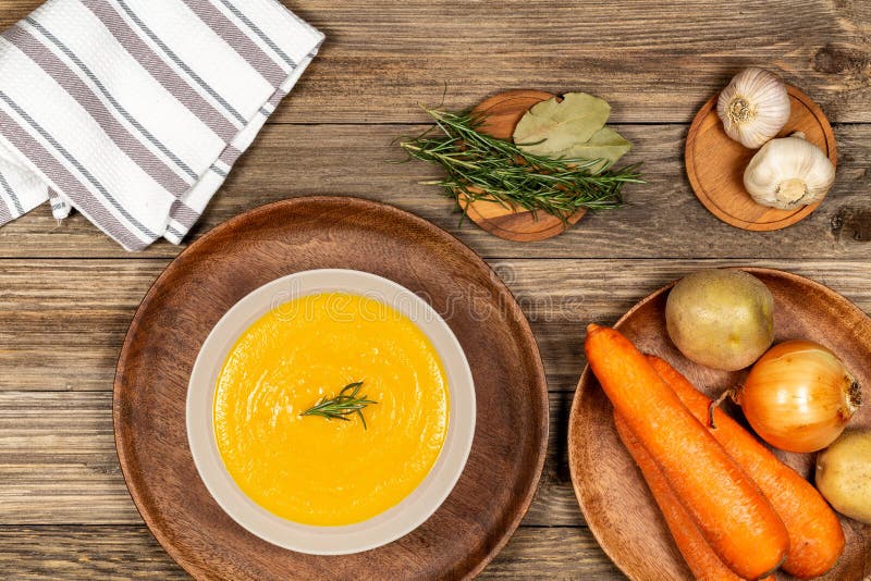 Carrot and potato soup bowl on a drift wood table with vegetables in a wood plate, napkin, garlic, laurel and rosemary. Flat lay stock image Carrot and potato soup bowl on a drift wood table with vegetables in a wood plate, napkin, garlic, laurel and rosemary. Flat lay stock image