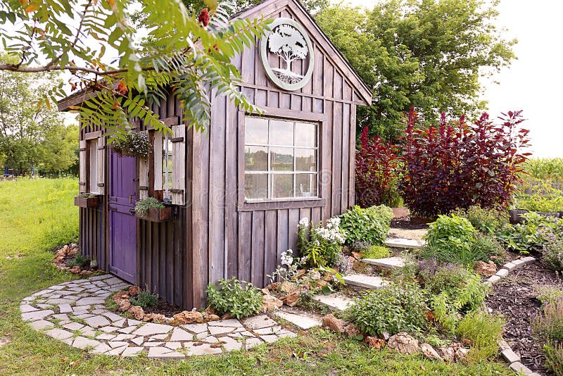 A custom built cottage like garden shed with a cobblestone sidewalk, purple door, window flower boxes, and wood board and batten siding. A custom built cottage like garden shed with a cobblestone sidewalk, purple door, window flower boxes, and wood board and batten siding