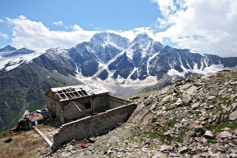 Old disturbed house among stones in Caucasus mountains. At the background there are snow-covered rocks, blue sky and white clouds. Elbrus area, near peak Cheget. Old disturbed house among stones in Caucasus mountains. At the background there are snow-covered rocks, blue sky and white clouds. Elbrus area, near peak Cheget.