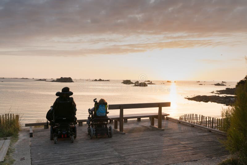 Casal deficiente em cadeiras de rodas desfrutam de um belo pôr do sol do oceano junto de um ponto de vista de acesso à praia defi imagens de stock royalty free Casal deficiente em cadeiras de rodas desfrutam de um belo pôr do sol do oceano junto de um ponto de vista de acesso à praia defi imagens de stock royalty free