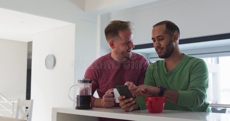 Casal gay multiétnico na cozinha falando um usando smartphone